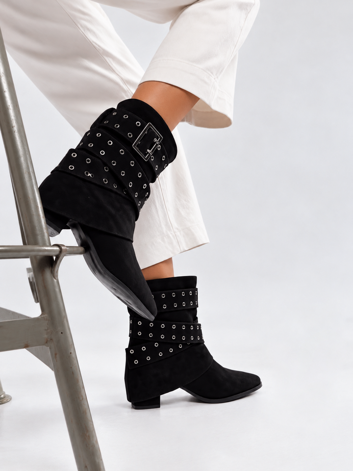 Black faux suede buckle strap slouch ankle boot – worn on model, seated on a stool
