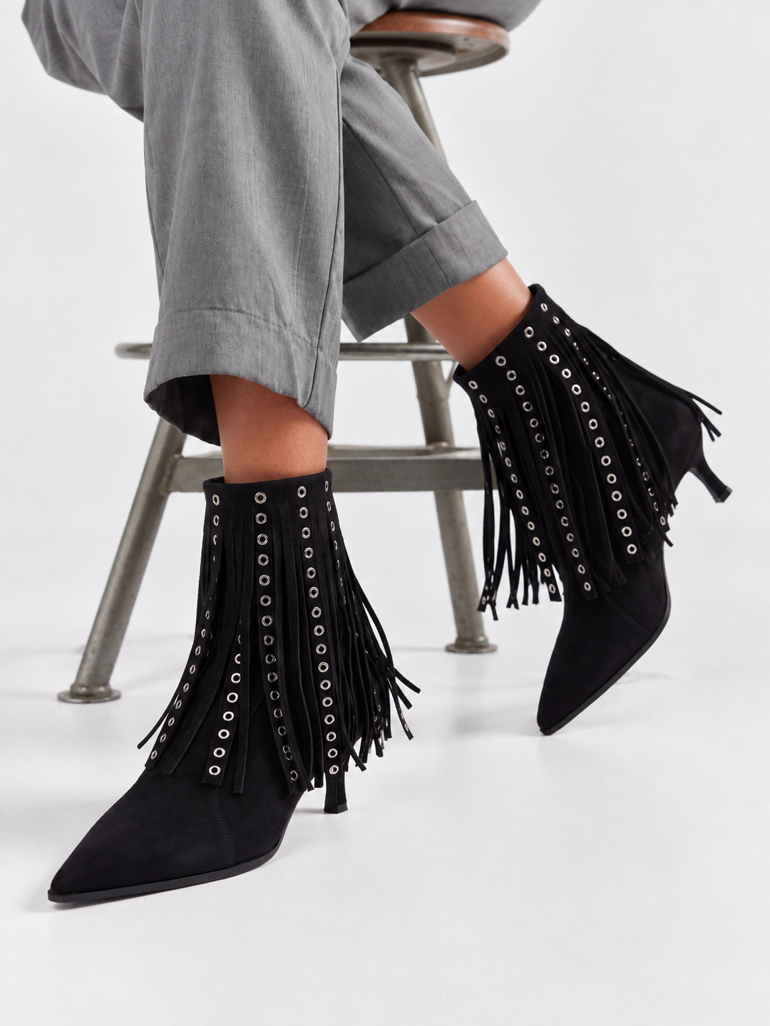 Black faux suede fringe heeled ankle boot – worn on model, seated, front close-up